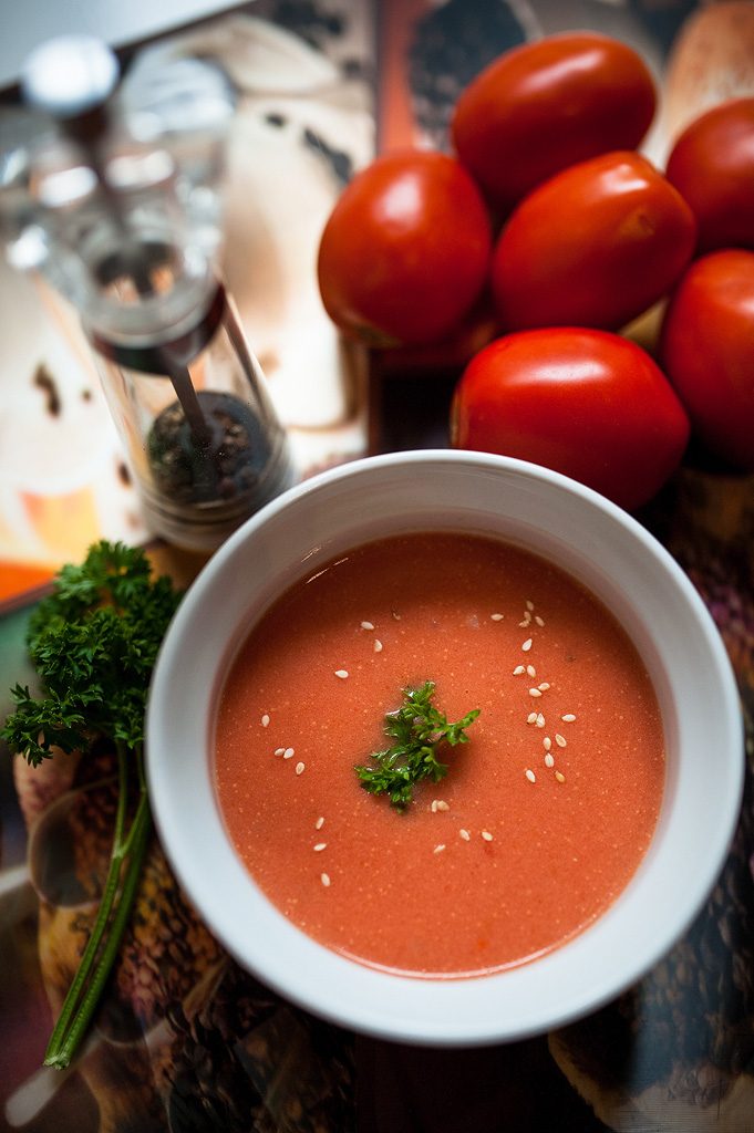 A bowl of tomato shorba topped with coriander and sesame, tomatoes behind